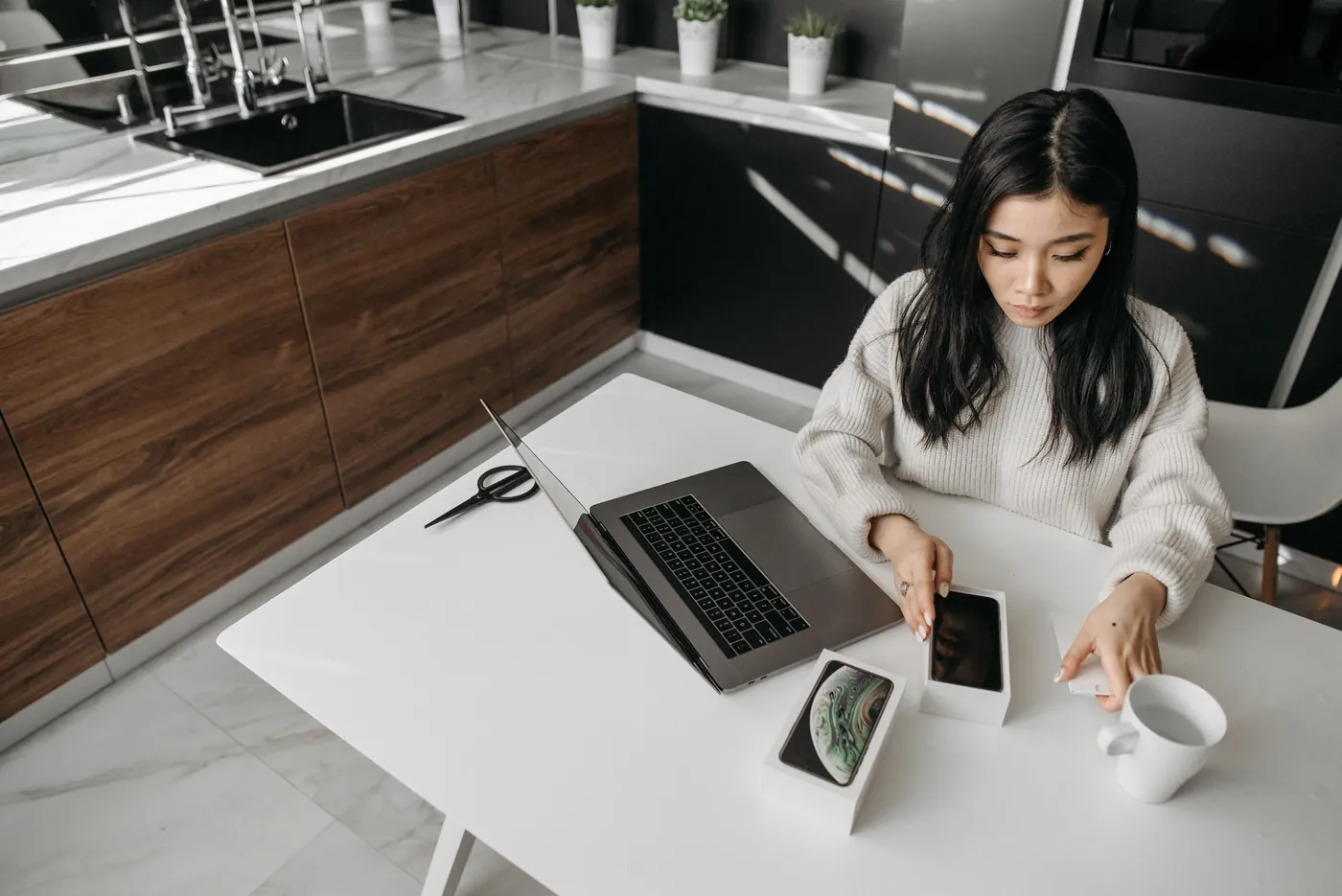 A woman unboxing a phone beside an open laptop at a kitchen table.