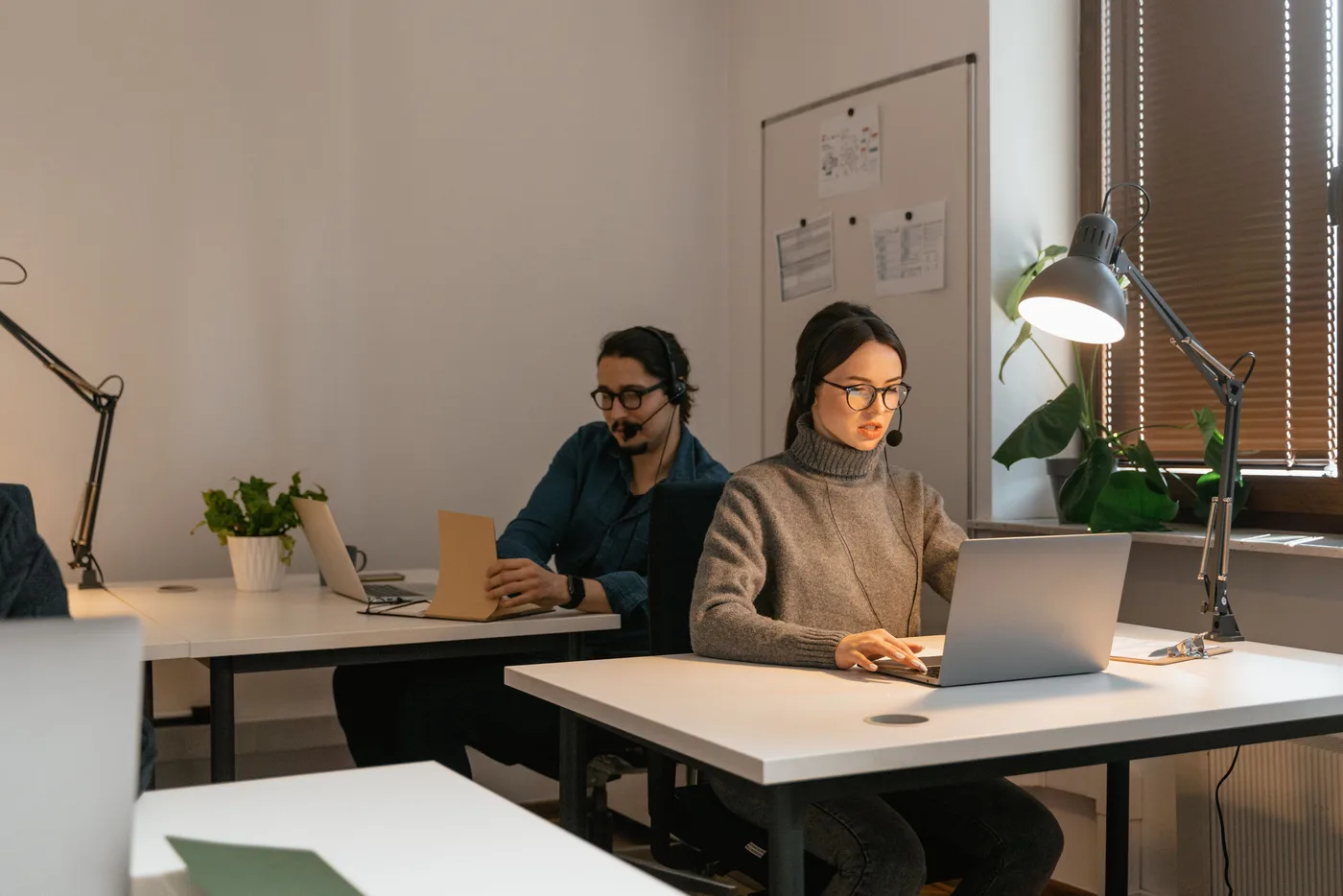 Two colleagues wearing headsets work with laptops and notes in a shared office.